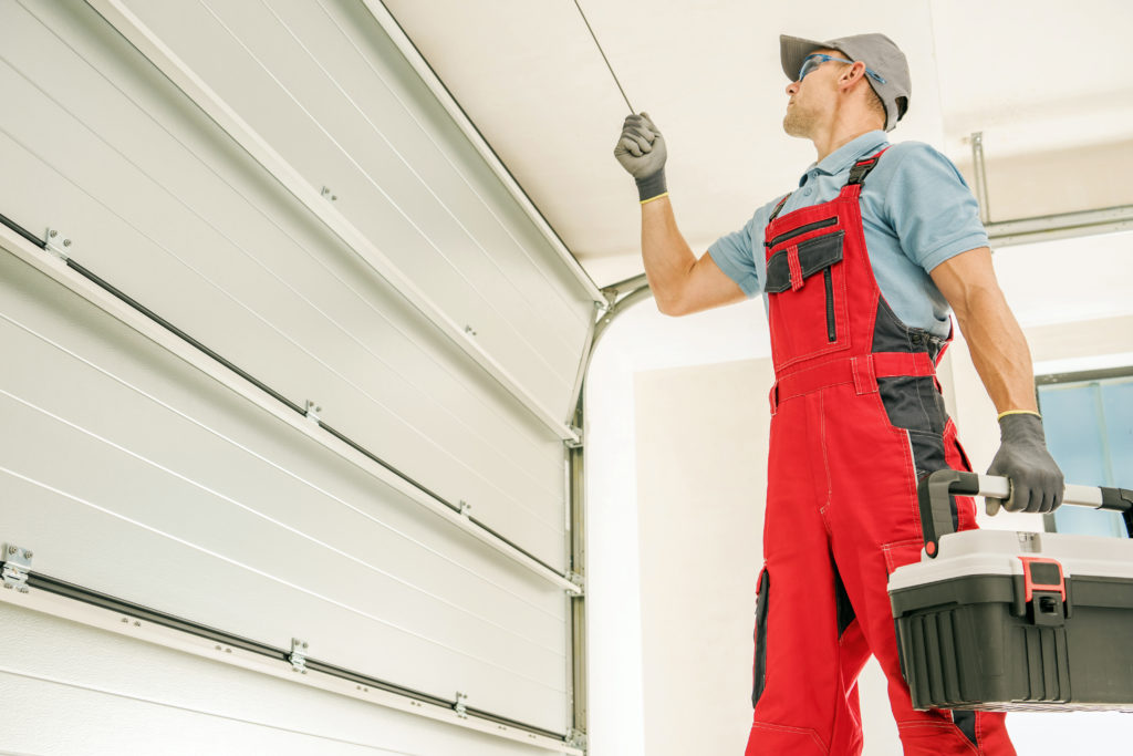 A technician from the garage door company, dressed in red coveralls and a cap, adjusts the garage door mechanism while holding a toolbox.